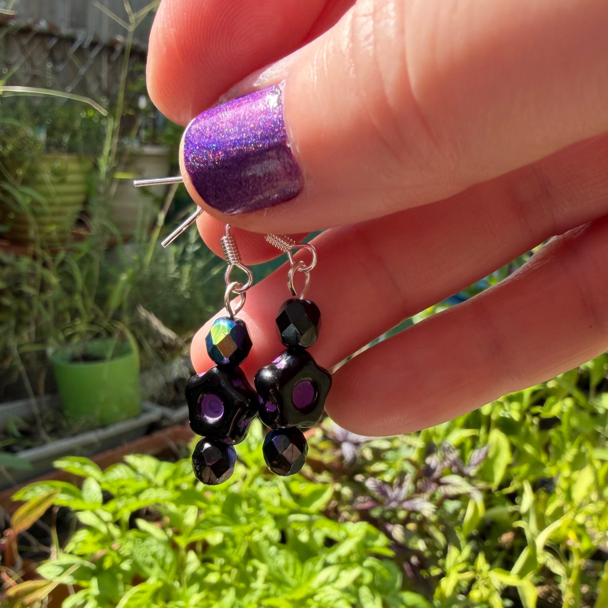 A close-up of a hand with purple glitter nail polish holding a pair of dangling earrings with black faceted beads and silver hooks, set against a blurred green garden background.