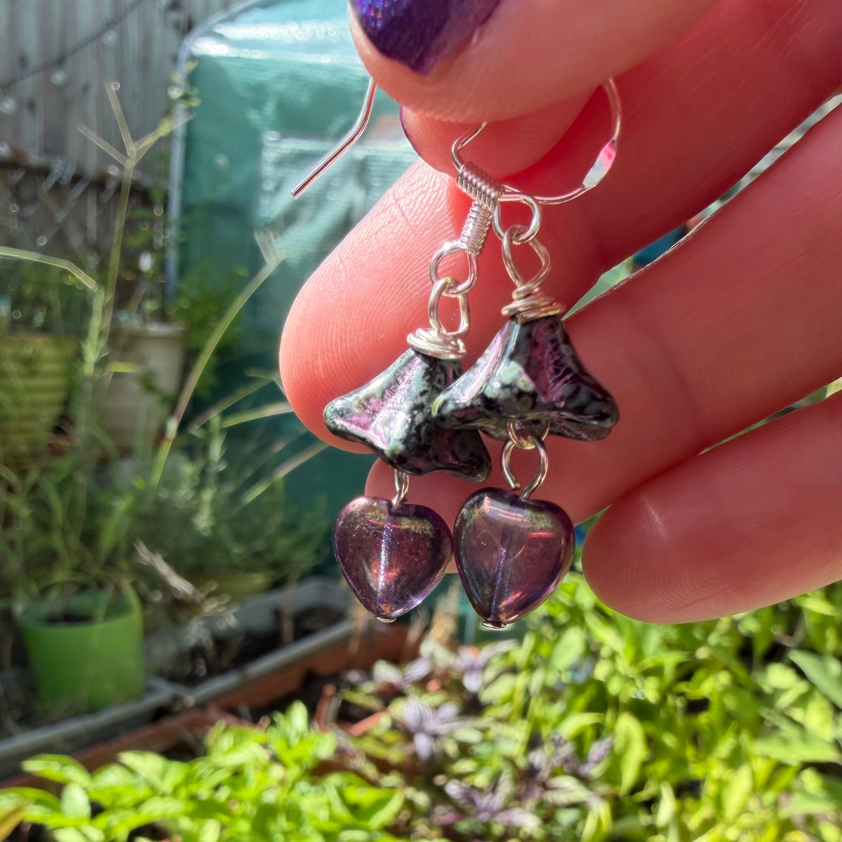 A close-up of a hand holding a pair of dangling earrings with silver-toned hooks and dark purple heart-shaped beads, set against a blurred outdoor garden background.