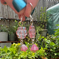 Pink flower and bell dangle earrings held up by a hand in front of a garden bed with potted plants.