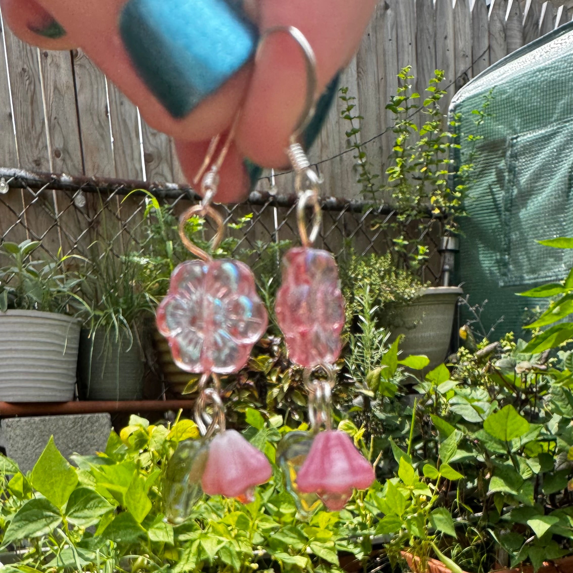Pink flower and bell dangle earrings held up by a hand in front of a garden bed with potted plants.