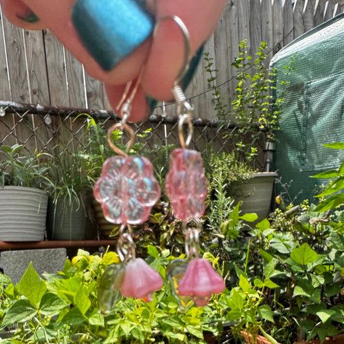 Pink flower and bell dangle earrings held up by a hand in front of a garden bed with potted plants.
