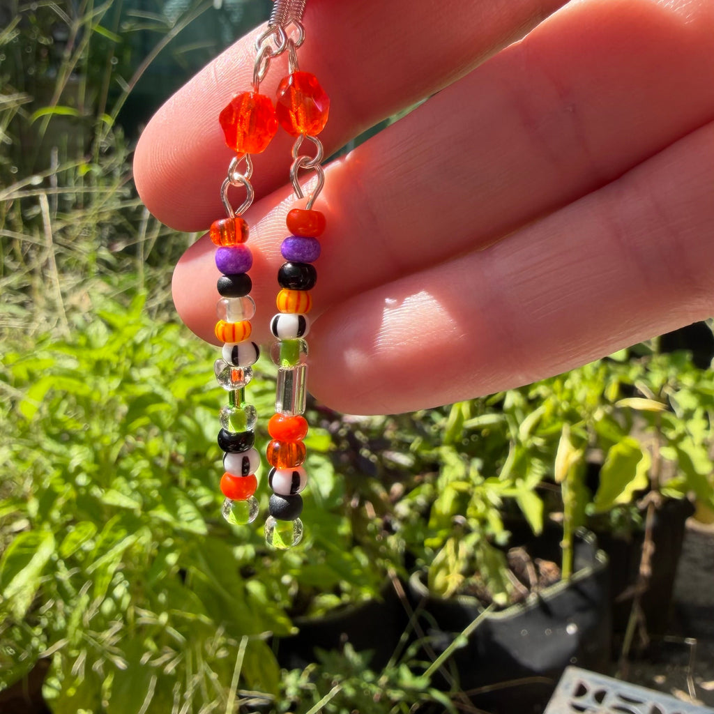 A close-up of a hand holding a pair of dangling beaded earrings outdoors. The earrings feature clear and silver-toned beads with orange, purple, black, white, and red accents.