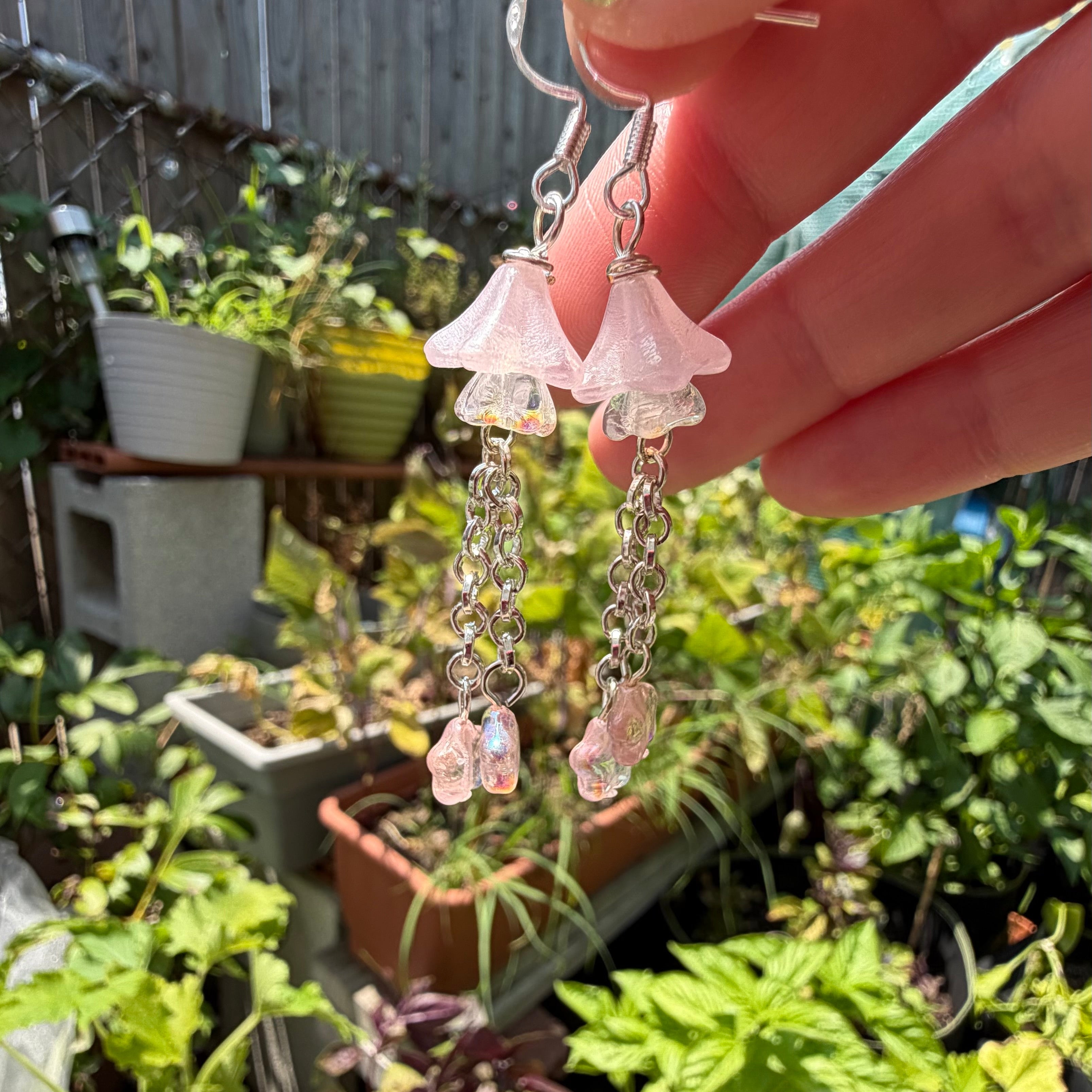 A pair of dangling earrings held by a hand outdoors in a garden setting. The earrings feature light pink bell-shaped beads, silver-toned chain links, and small pink flower charms.