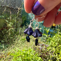 A close-up of a hand with purple glitter nail polish holding a pair of dangling earrings outdoors in a garden. The earrings have silver hooks and silver wire loops, with dark blue to black iridescent bell-shaped beads hanging below.