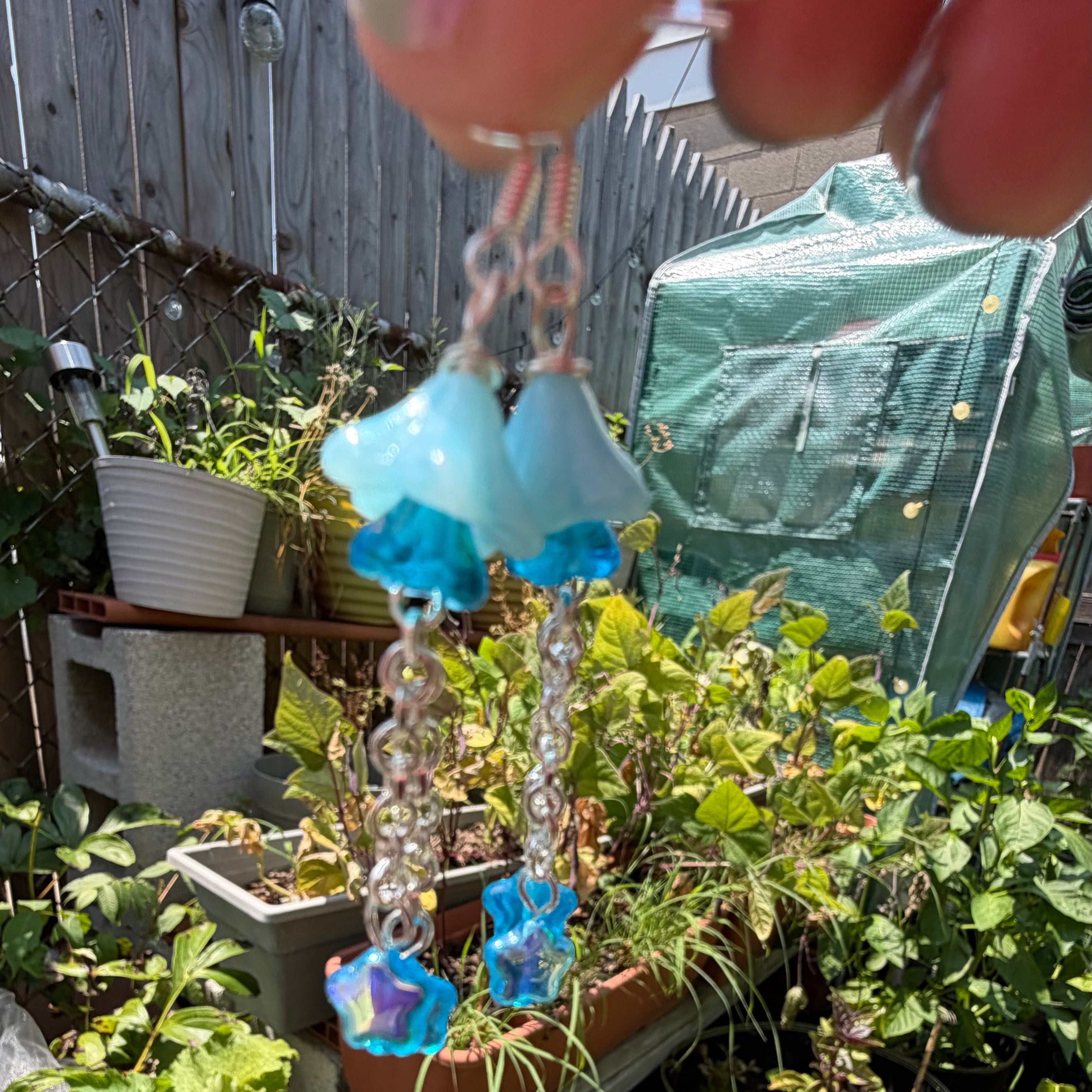 A pair of dangling earrings being held by a hand in a garden. The earrings feature light blue flower-shaped beads, iridescent blue and purple beads, and silver-toned chain links.