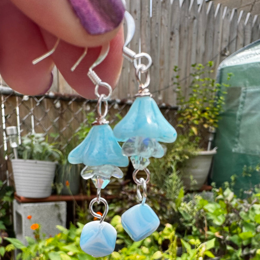 A pair of dangling earrings being held by a hand, featuring light blue bell-shaped beads and faceted blue beads on silver hooks, set against a background of a garden with potted plants.