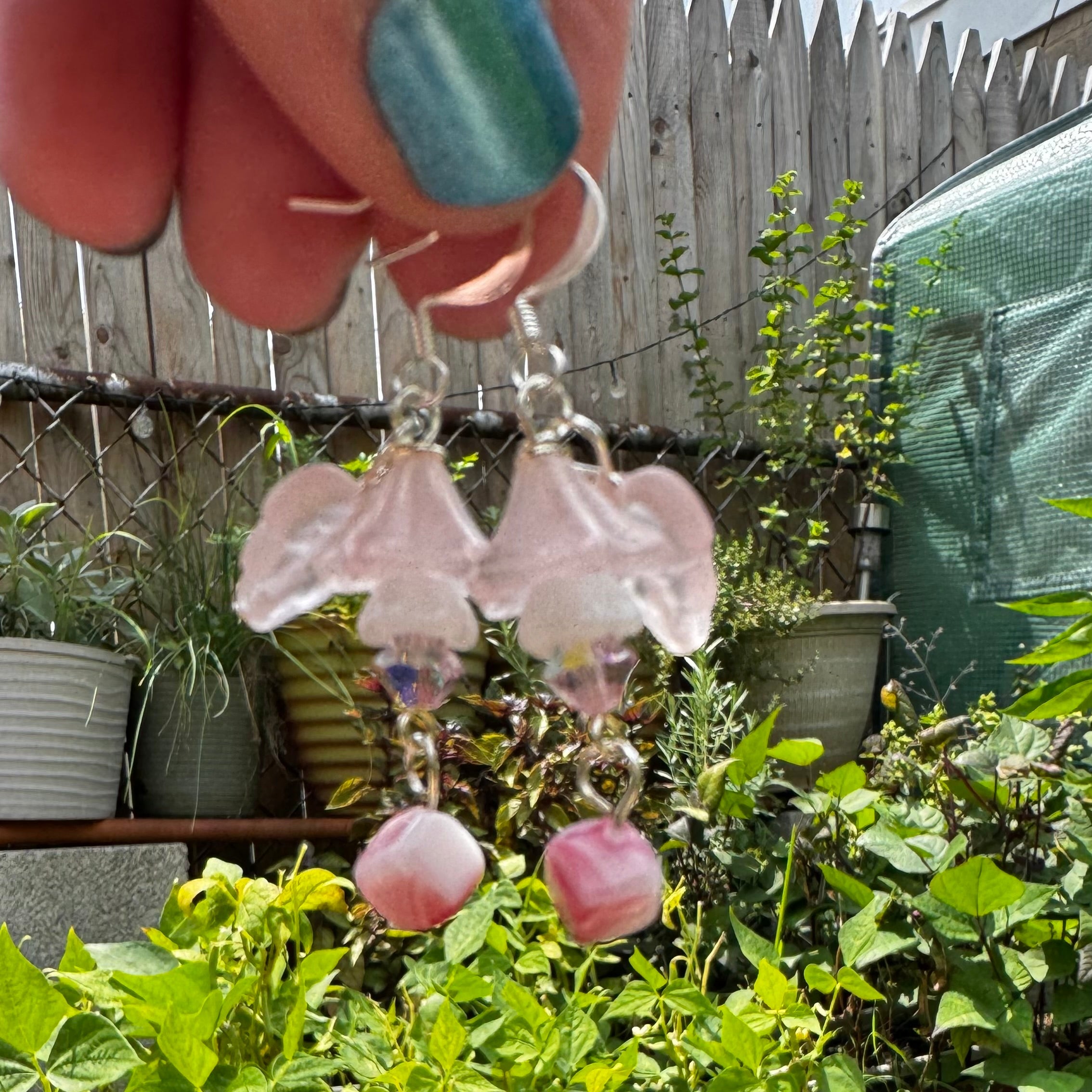 Pink bell-shaped flower dangle earrings with silver-toned hooks, held up by a hand in front of a garden bed with potted green plants.