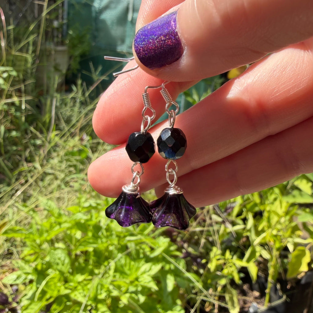 A close-up of a hand holding a pair of handmade dangle earrings outdoors. The earrings feature faceted black beads and purple flower-shaped beads with silver hooks.