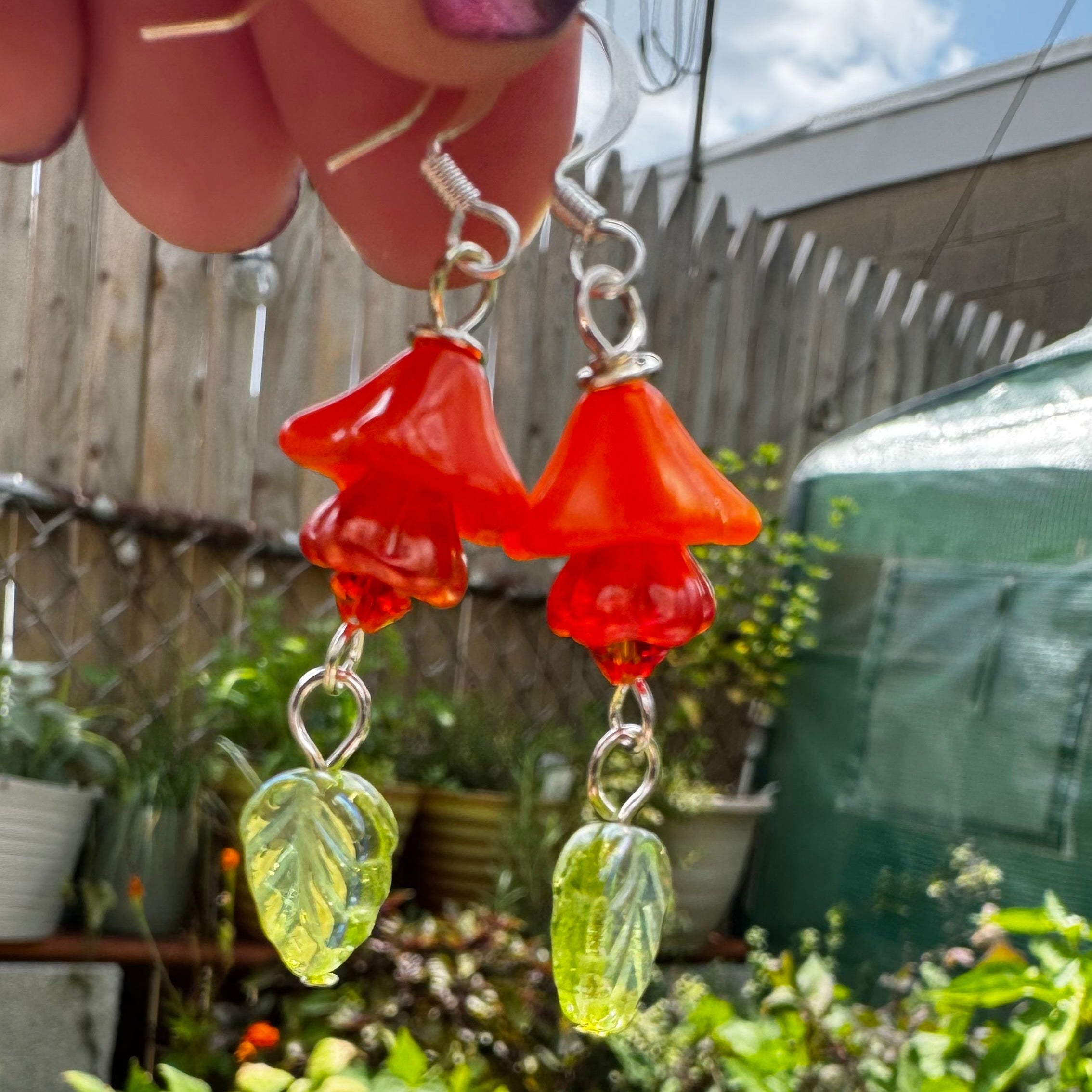 A close-up of handmade dangle earrings held by a hand, featuring translucent orange mushroom-shaped beads and green leaf-shaped beads, set against a blurred garden background.