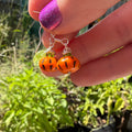 A close-up of a hand with glittery purple nail polish holding a pair of orange pumpkin-shaped drop earrings with black facial features and silver hooks, set against a blurred green garden background.