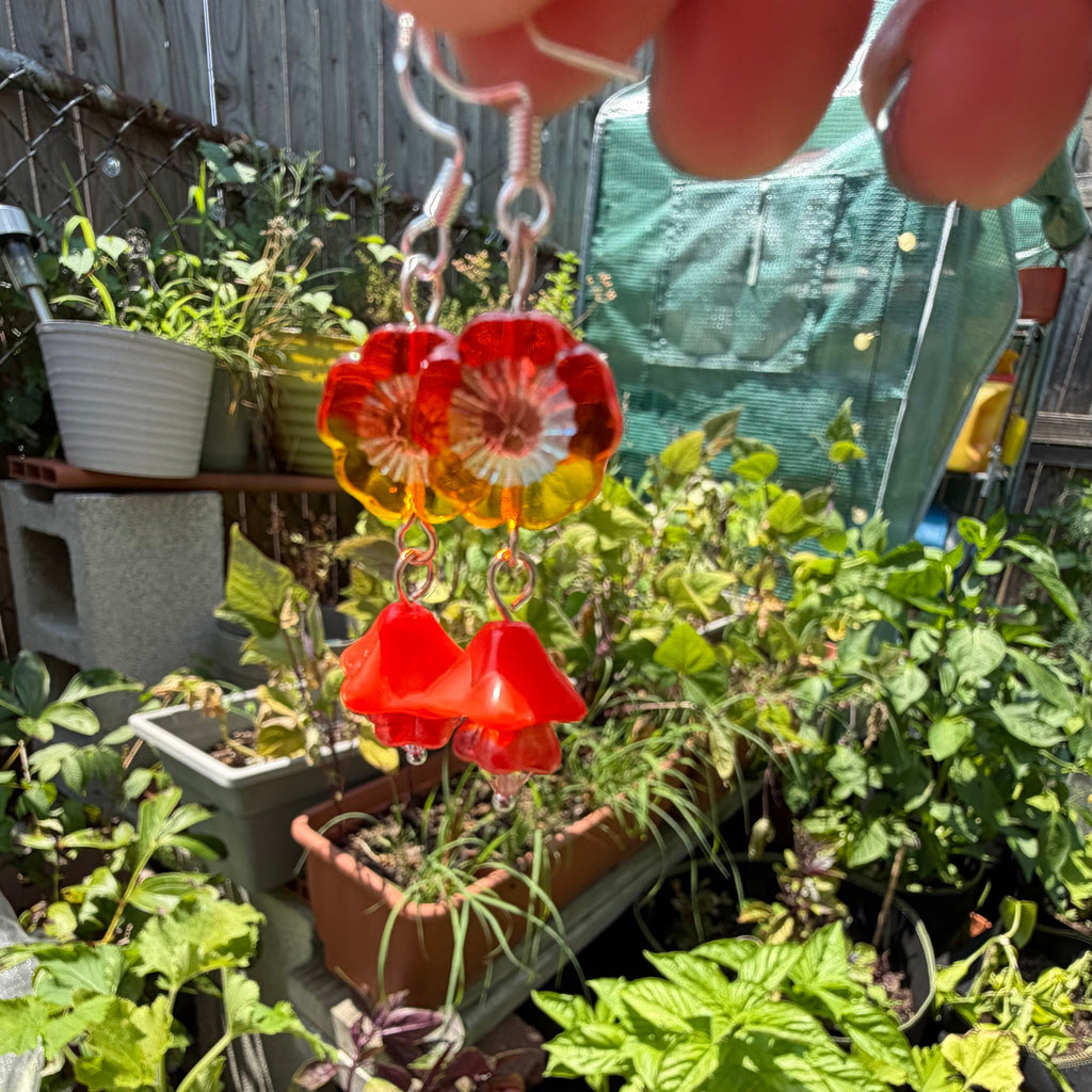 A close-up of a hand holding a pair of dangling earrings with flower-shaped charms in red, orange, and yellow, set against a busy garden background with plants and pots.