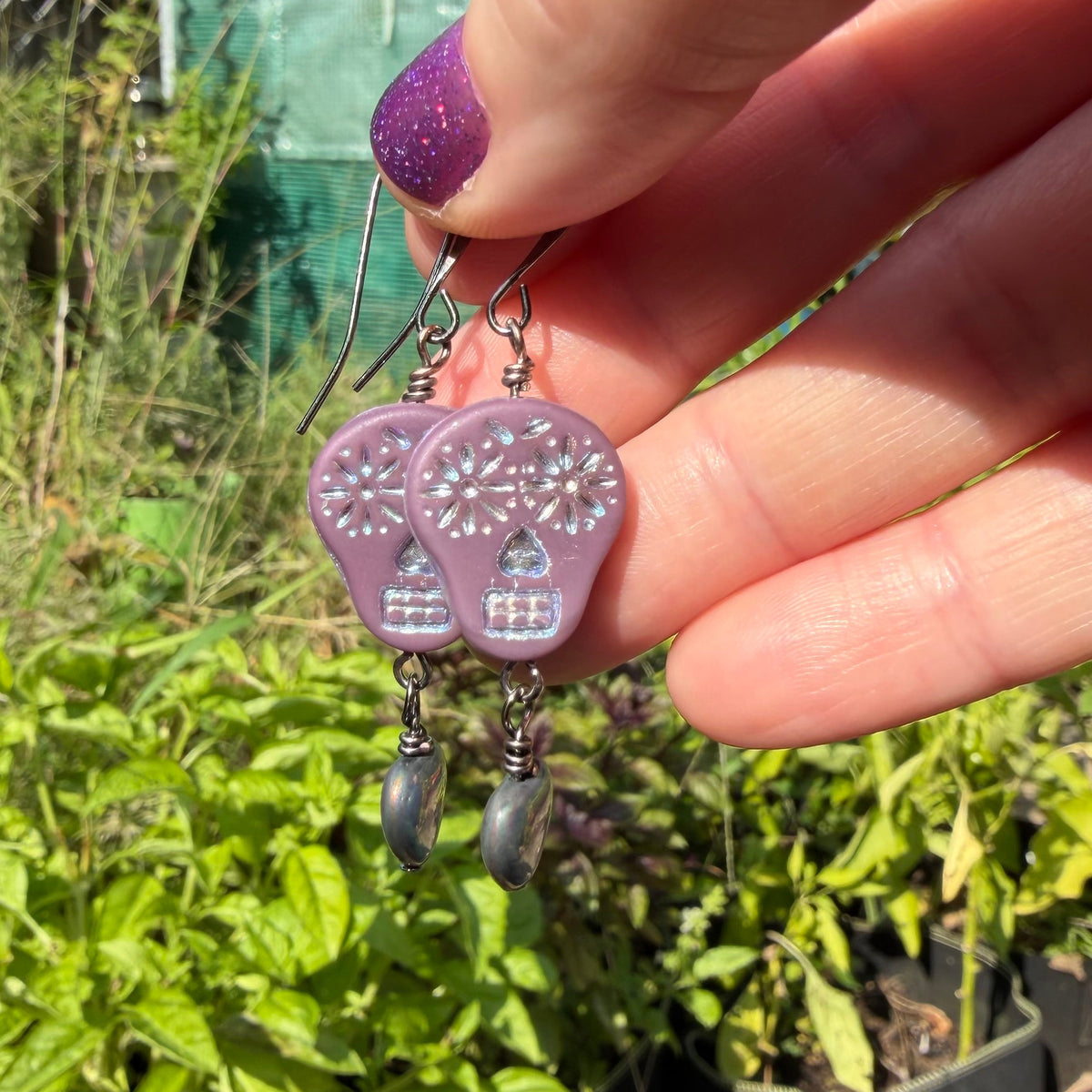 A hand with purple glitter nail polish holds a pair of dangling earrings outdoors in front of green plants. The earrings feature lavender-purple mushroom charms with white floral details and dark gray bead drops.
