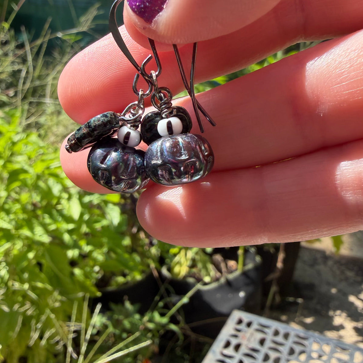 A close-up of a hand holding a pair of dangling earrings with dark beads and small white-and-black eye-like charms, photographed outdoors with a garden background.