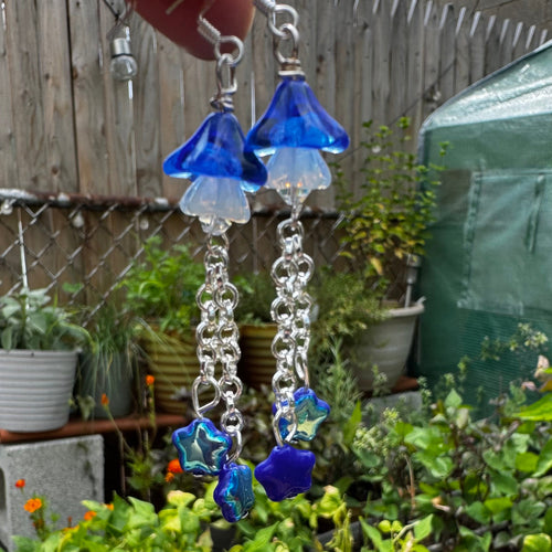 A pair of handmade dangle earrings held by a hand, featuring blue and clear glass flower charms, silver chain links, and blue star-shaped beads, set against a background of potted plants and a wooden fence.