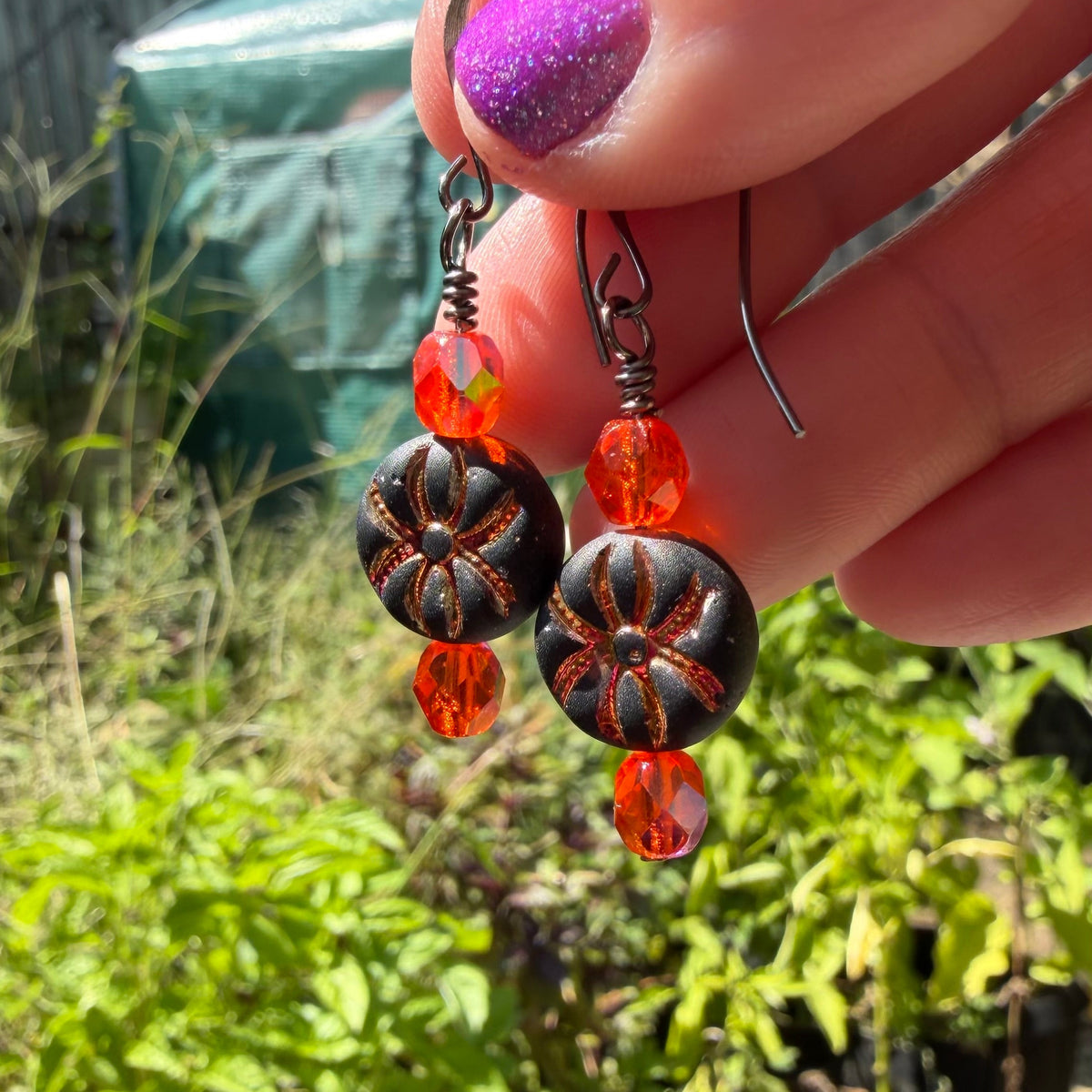 A close-up of a hand holding a pair of handmade dangle earrings with orange faceted beads, dark brown patterned round beads, and silver-toned hooks, set against a blurred garden background.