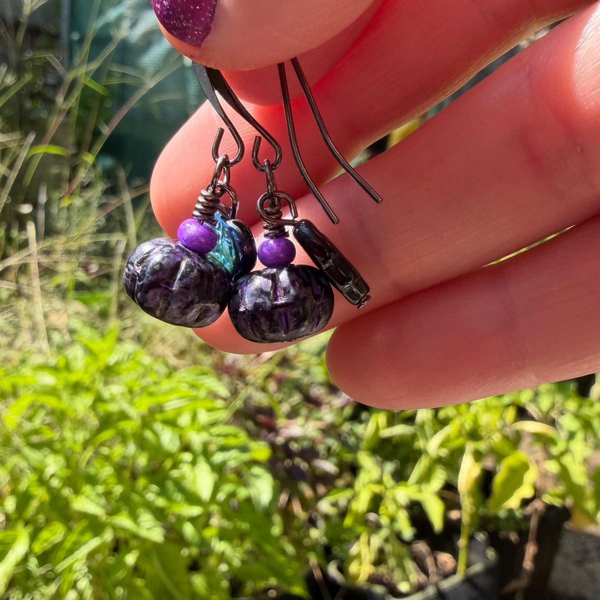 A close-up of a hand holding a pair of handmade dangle earrings with dark purple marbled beads, small purple and teal beads, and dark metal hooks, set against a blurry garden background.