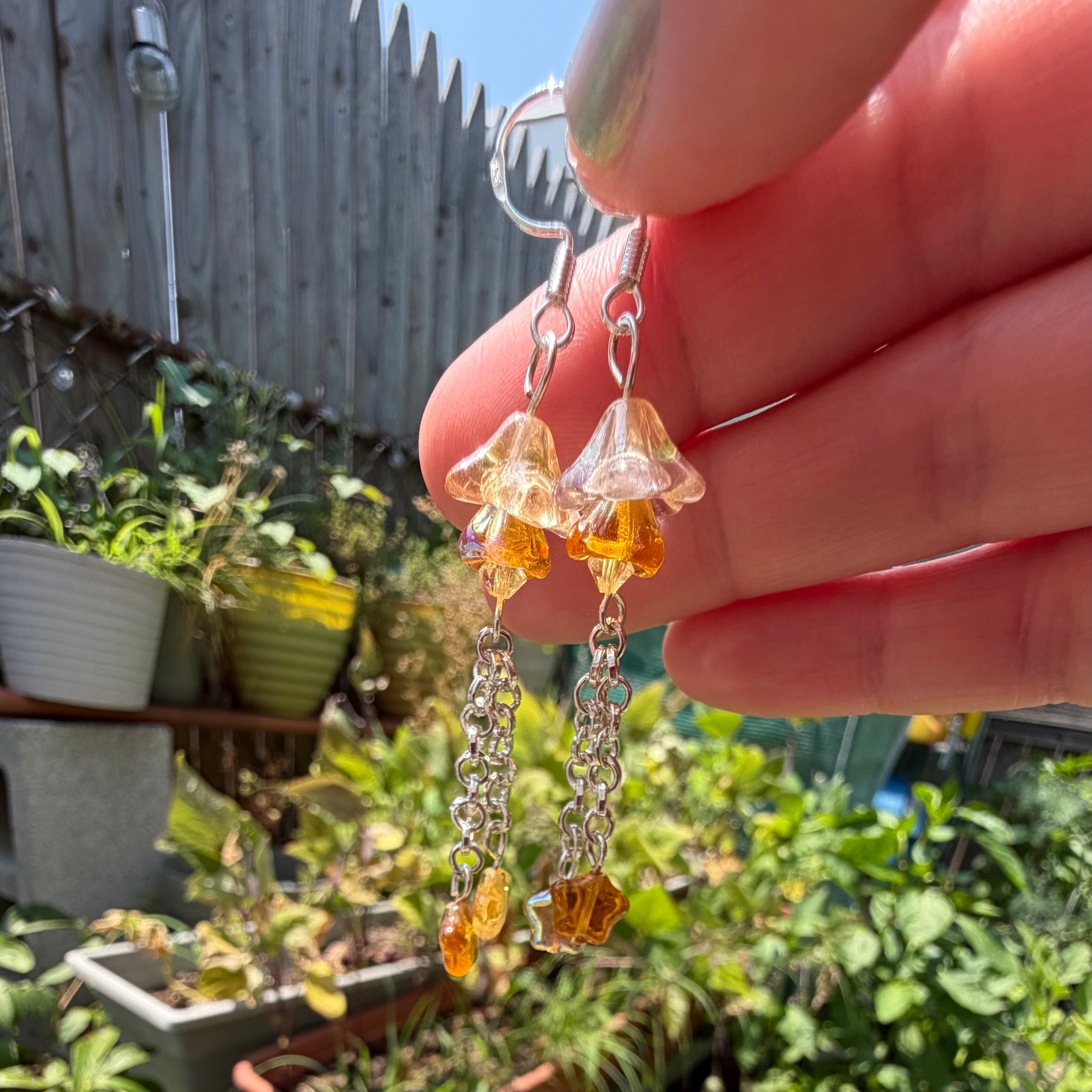 A person's hand holding up a pair of dangling earrings with clear bell-shaped crystals, amber-colored beads, and silver-toned chain drops, set against a blurred outdoor garden background.