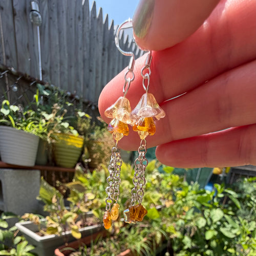 A person's hand holding up a pair of dangling earrings with clear bell-shaped crystals, amber-colored beads, and silver-toned chain drops, set against a blurred outdoor garden background.