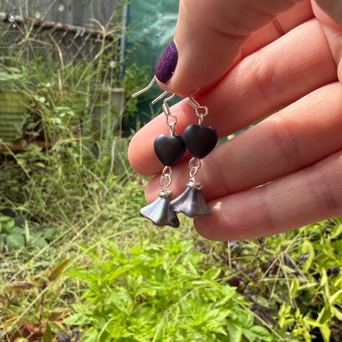 A close-up of a hand holding a pair of dangle earrings with black heart-shaped beads and silver bell-shaped charms, set against a blurred garden background.