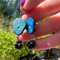 A close-up of a hand holding a pair of sugar skull and black rose dangle earrings outdoors, with green plants and a metal garden bench in the background.