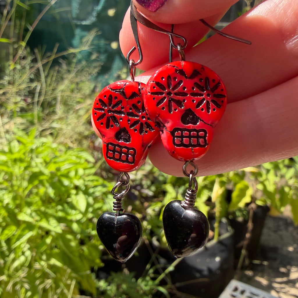 A close-up of a hand with purple glitter nail polish holding a pair of red sugar skull dangle earrings with black heart charms, set against a blurry outdoor garden background.