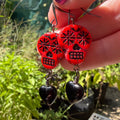 A close-up of a hand with purple glitter nail polish holding a pair of red sugar skull dangle earrings with black heart charms, set against a blurry outdoor garden background.