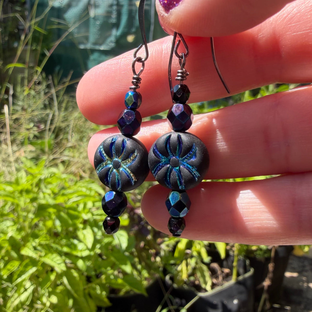 A close-up of a hand holding a pair of handmade drop earrings with dark blue and black patterned beads and dark metal hooks, set against a blurred outdoor garden background.