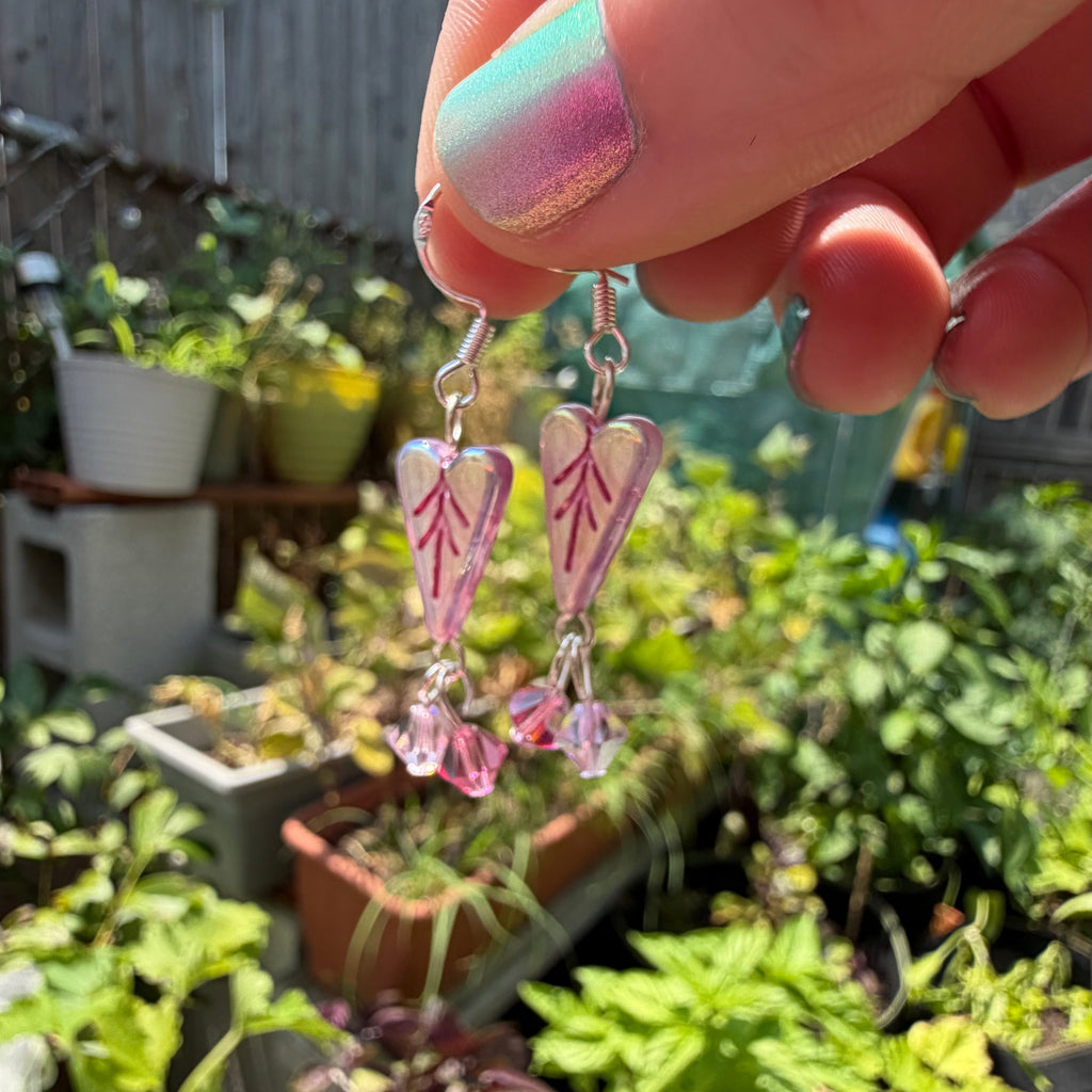 A hand with pastel pink and mint nail polish holds a pair of dangling earrings with pink and white leaf-shaped charms and silver hooks against a blurred garden background.