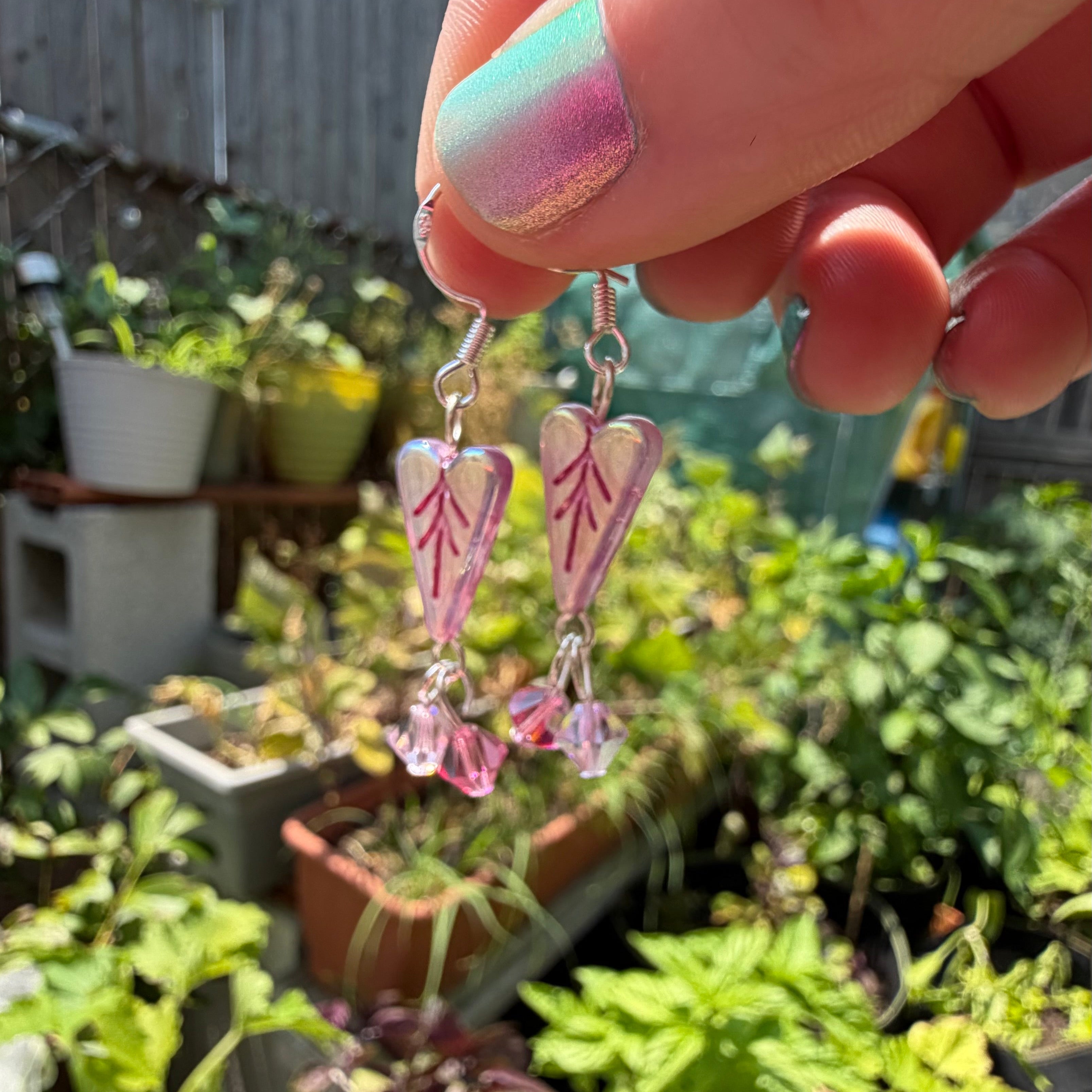 A hand with pastel pink and mint nail polish holds a pair of dangling earrings with pink and white leaf-shaped charms and silver hooks against a blurred garden background.