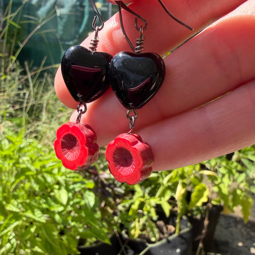 A close-up of a hand holding a pair of black heart-shaped dangle earrings with red flower-shaped charms, set against a blurred outdoor garden background.
