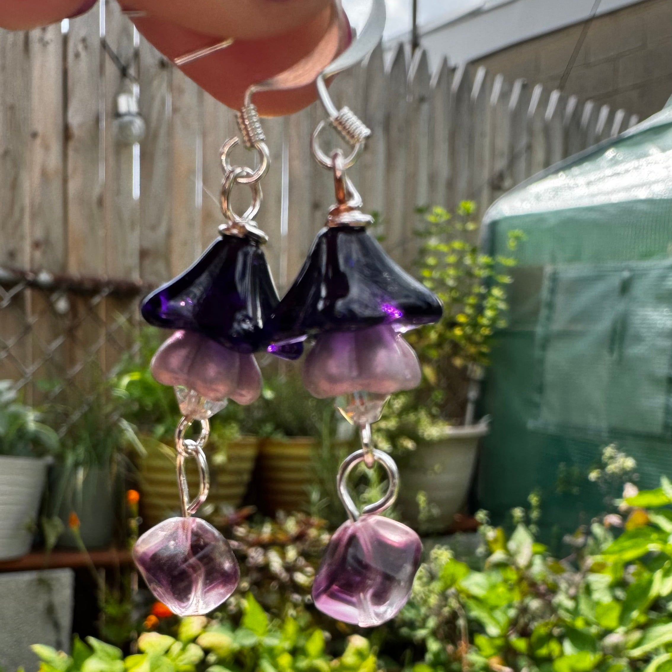 A pair of handmade dangle earrings held by a hand, featuring dark purple and light purple beads with silver hooks, set against a background of potted green plants and a wooden fence.