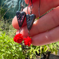A close-up of a hand holding a pair of gothic-style dangle earrings outdoors. The earrings feature dark, textured heart-shaped charms and small red rose charms dangling below, set against a blurred garden background.