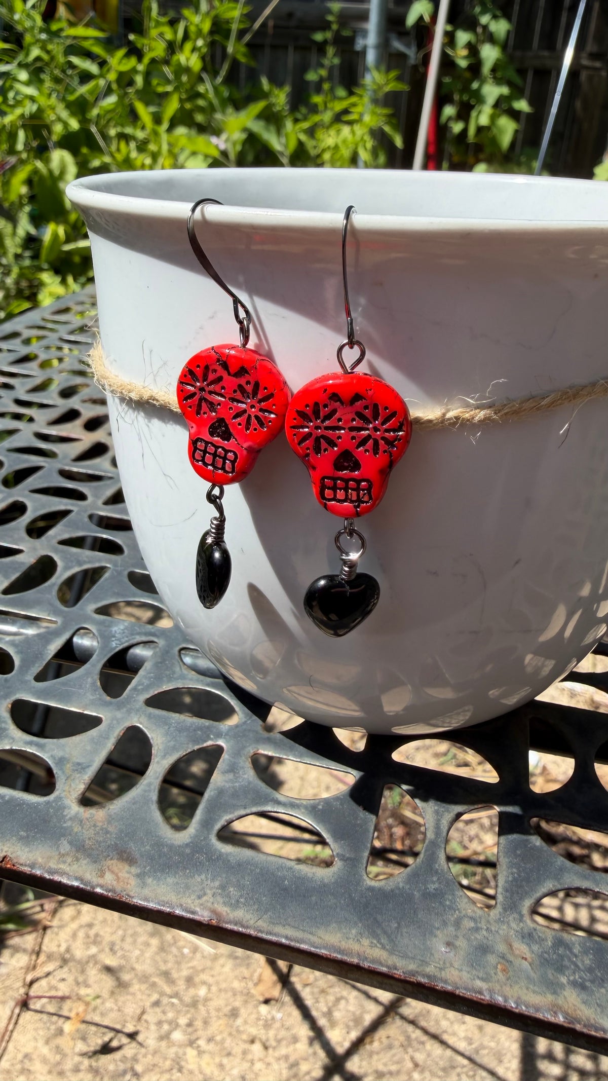 A pair of red sugar skull drop earrings with black heart charms hanging from a white planter on an outdoor metal table.