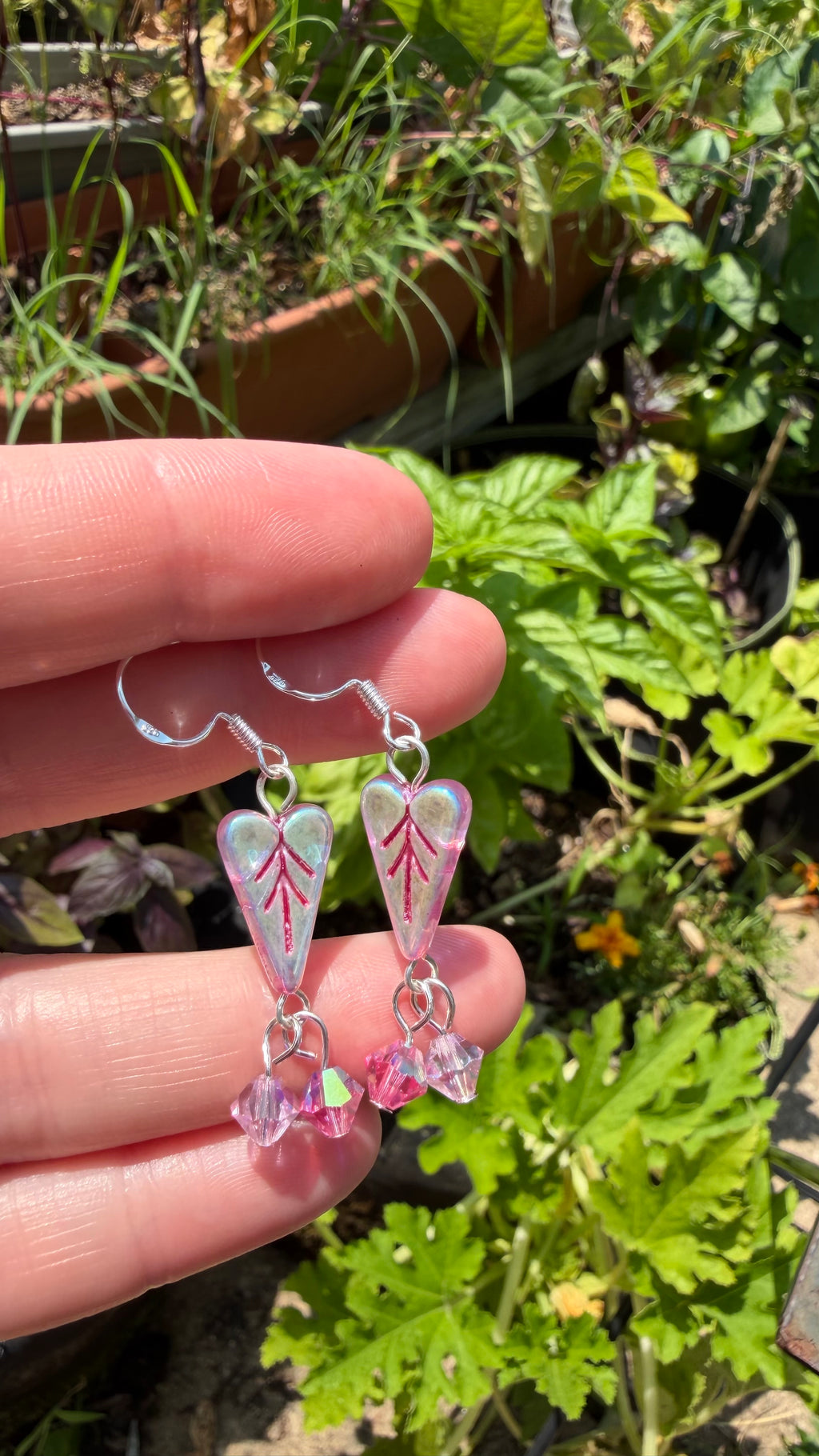 A hand holding a pair of dangling earrings outdoors, with green plants and garden foliage blurred in the background. The earrings have silver fishhook wires, iridescent pink heart-shaped beads with red vein-like lines, and dangling pink faceted beads.