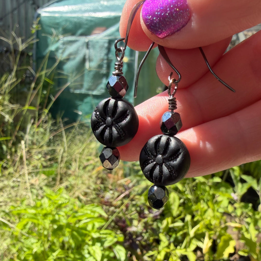 A close-up of a hand with glittery purple nail polish holding a pair of black beaded dangle earrings outdoors in a garden setting.