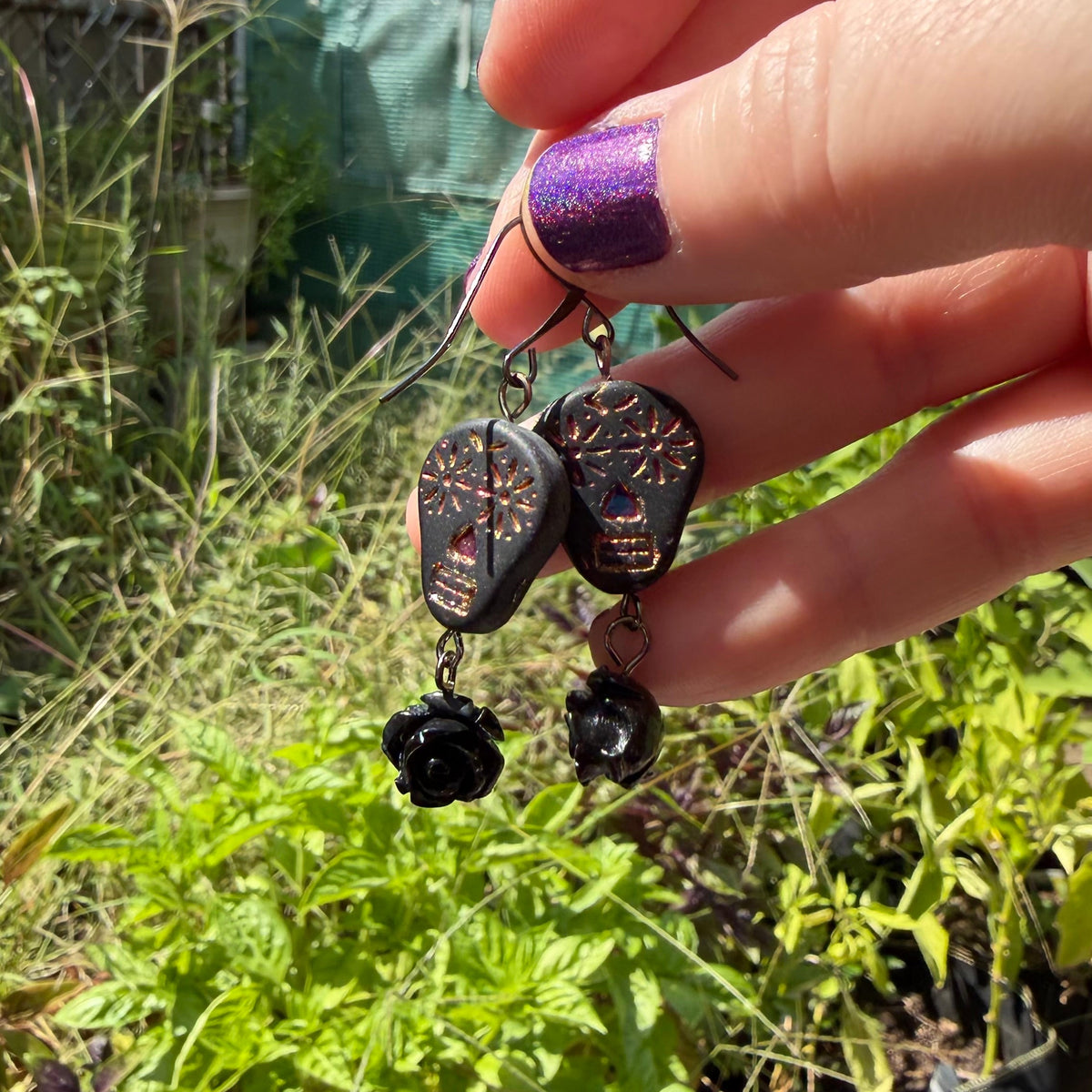 A hand with purple glitter nail polish holds a pair of black dangle earrings featuring carved guitar-pick-shaped charms and small black beads, set against a blurry outdoor garden background.