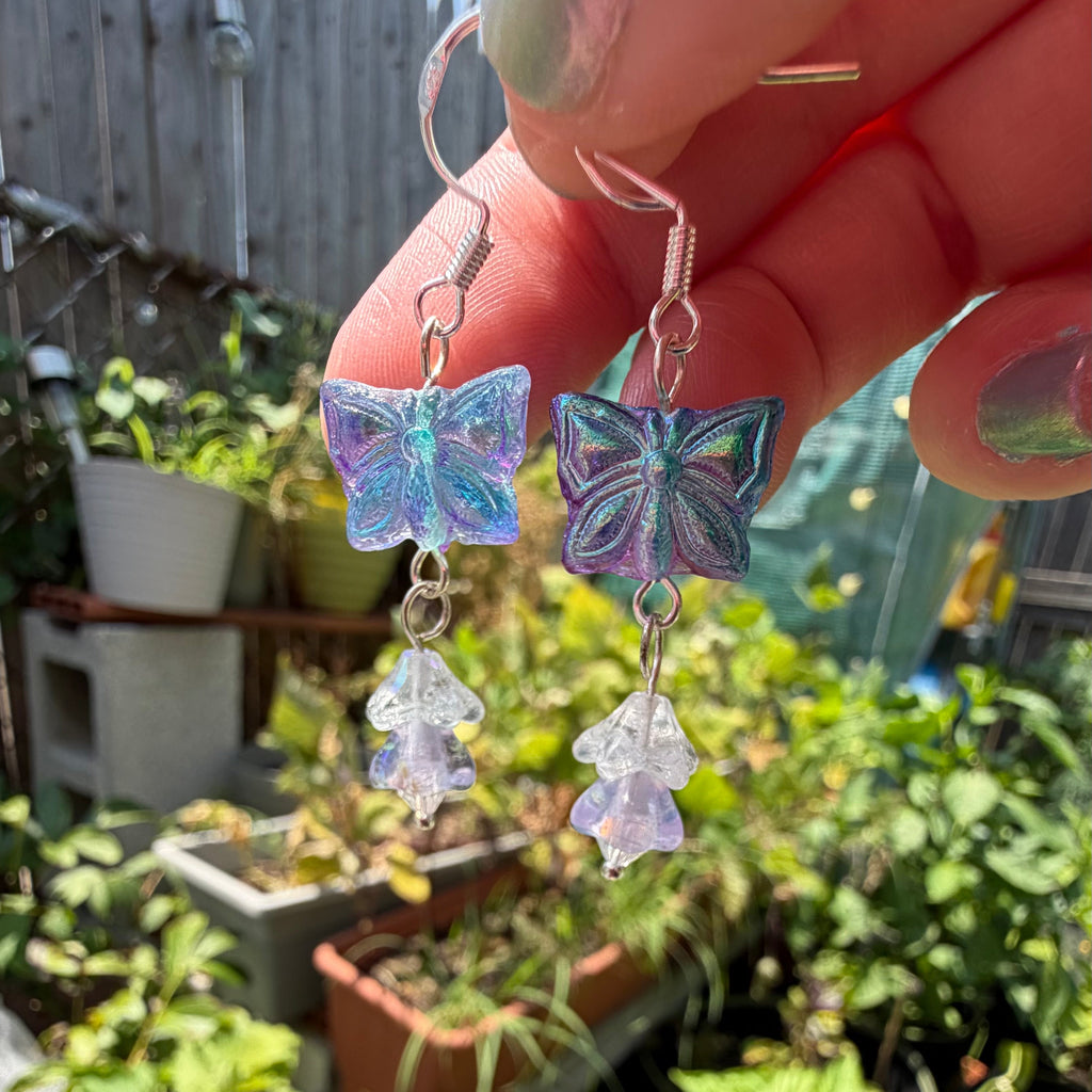 A close-up of a hand holding a pair of dangling earrings outdoors in front of a garden. The earrings have iridescent blue and purple butterfly charms with clear flower-shaped drops.