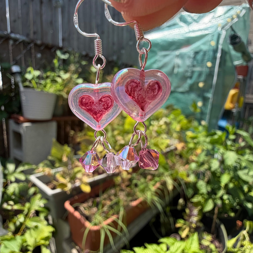 A pair of dangling heart-shaped earrings with pink glitter and clear rhinestone charms, held by a hand in an outdoor garden setting.