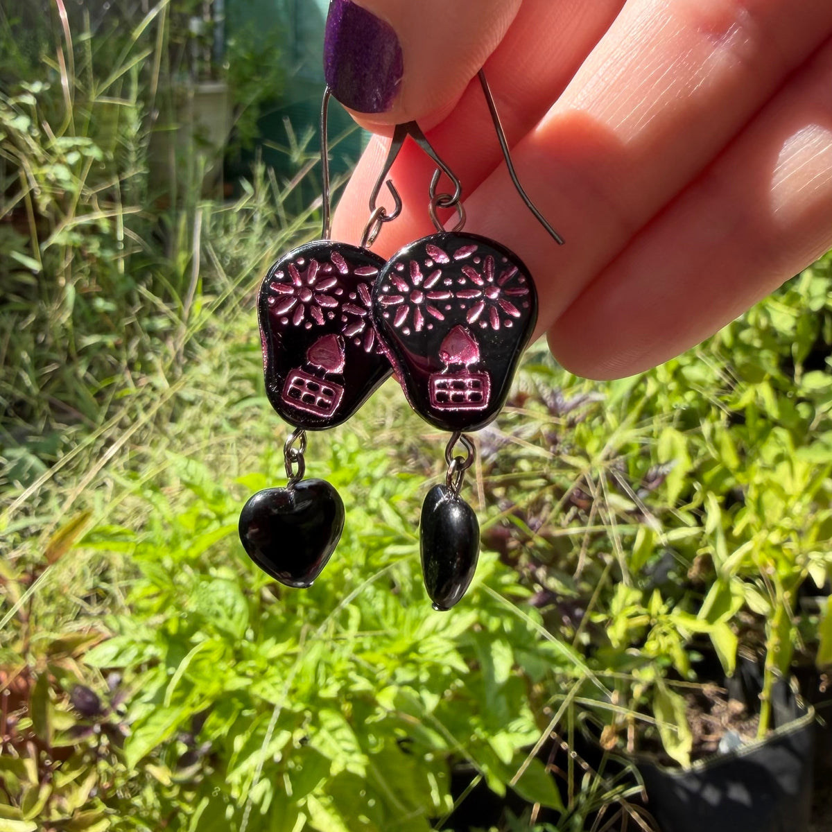 A hand with purple nail polish holds a pair of sugar skull and heart dangle earrings outdoors in front of a garden background.