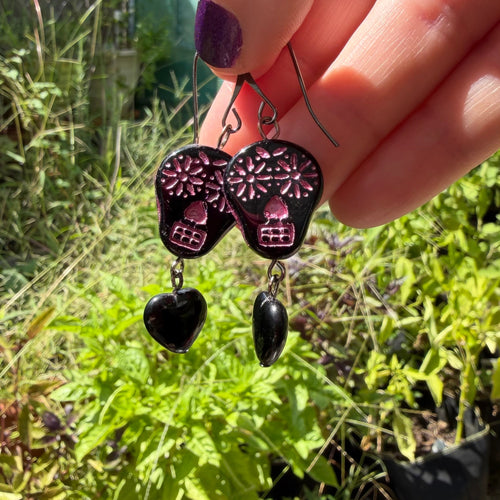 A hand with purple nail polish holds a pair of sugar skull and heart dangle earrings outdoors in front of a garden background.