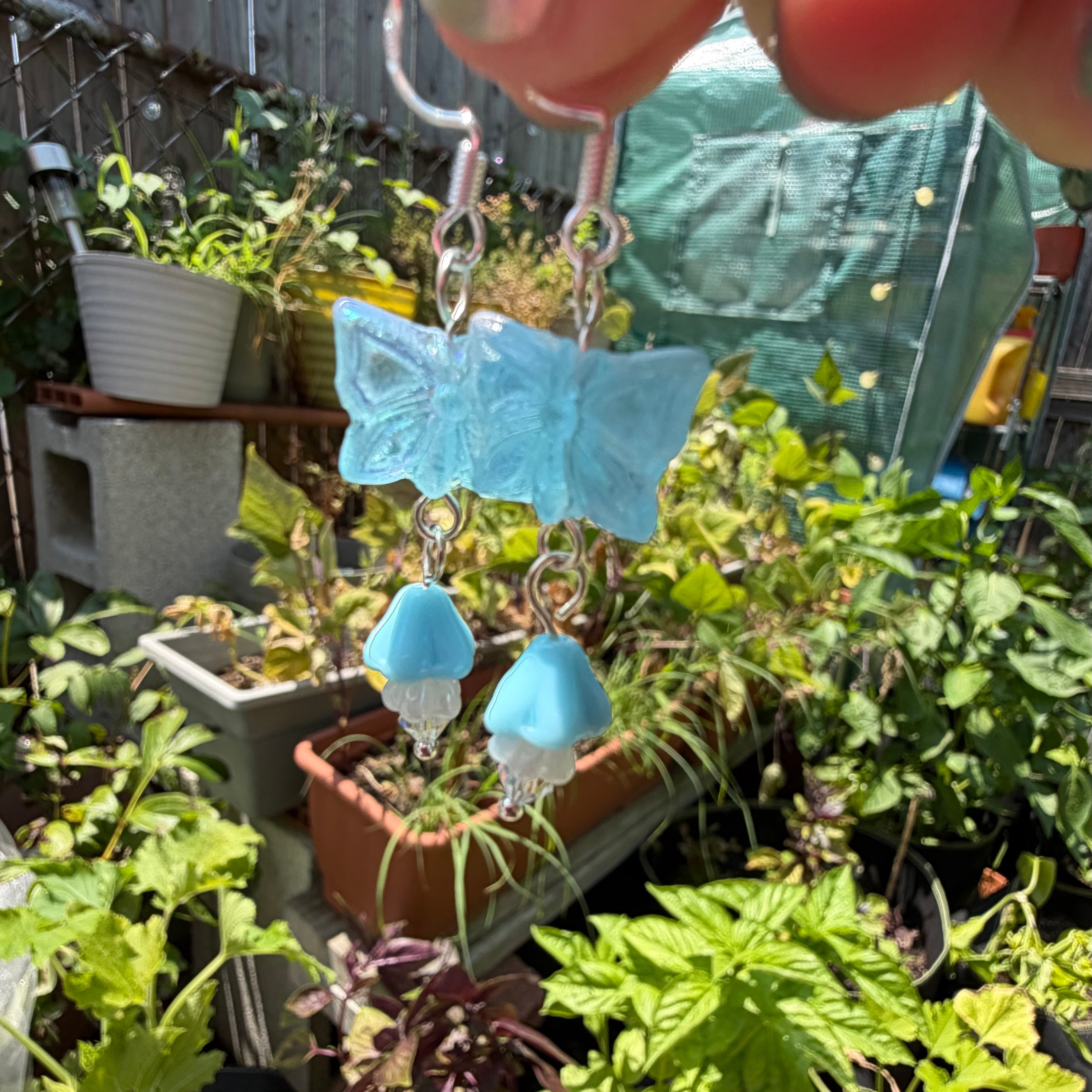 A close-up of a hand holding a pair of dangling earrings outdoors in a garden. The earrings feature a light blue butterfly-shaped charm with two small light blue bell-shaped charms below, attached to silver hooks.