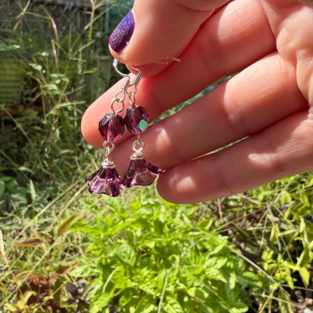 A hand holding a pair of dangling earrings with purple flower-shaped beads and silver hooks, photographed outdoors in a garden setting.